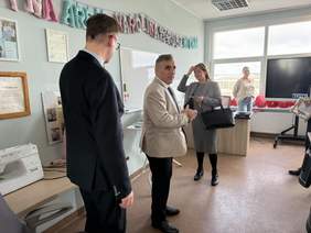 Pokaż zdjęcie: Kilka osób stoi w sali warsztatowej. Mężczyzna w jasnej marynarce rozmawia z innymi. W tle duże okno, monitor, biurko. Na ścianach wiszą certyfikaty i ozdoby. W rogu stoi maszyna do szycia.