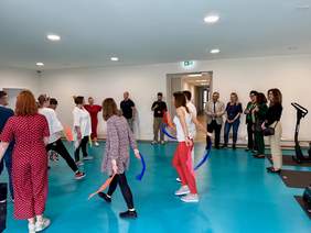 Pokaż zdjęcie: Osoby w sali gimnastycznej obserwują taniec lub ćwiczenia z chustami wykonywane przez grupę osób. Publiczność stoi przy ścianie, a ćwiczący w centrum w dynamicznych pozach.