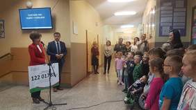 Pokaż zdjęcie: Na korytarzu szkolnym stoi kobieta i przemawia przez mikrofon do zgromadzonych dzieci i ich opiekunów. W reku trzyma dużą tablicę symbolizujaca czek.