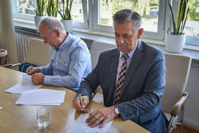 Dwóch mężczyzn siedzi przy stole, podpisują dokumenty