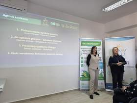 Na tle prezentacji stoja dwie kobiety, a jedna z nich przemawia