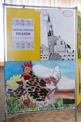 Pokaż zdjęcie: Na sztaludze wywieszone dwie prace plastyczna. Pierwsza to rysunek na którym są budynki w mieście. Drugi to malarstwo na którym są namalowane dwie kolorowe kury.