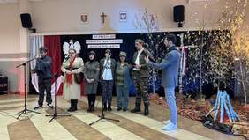 Pokaż zdjęcie: Grupa osób w strojach stylizowanych na wojsko występuje na scenie podczas XI Przeglądu Pieśni Patriotycznej „Jestem Polakiem” w Szumowie. W tle dekoracje z biało-czerwonych flag i symbolami narodowymi. Obok uczestników stoi osoba z gitarą, tworząc muzyczną oprawę występu.