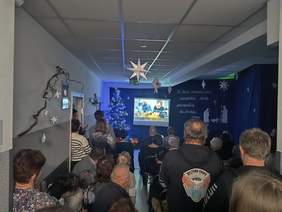 Pokaż zdjęcie: Zdjęcie z tyłu sali w kierunku ekranu, na którym wyświetlana jest prezentacja multimedialna.