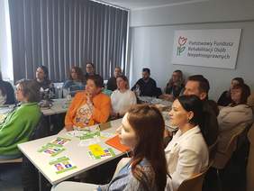 Pokaż zdjęcie: Grupa kilkunastu osób siedzących przy stole konferencyjnym w kształcie liter „U”, na których leżą kolorowe broszury, notesy oraz napoje. Przy stołach siedzi kilkanaście osób. Na prawej ścianie wisi prostokątna tablica z logo w kształcie tulipana i napisem: „Państwowy Fundusz Rehabilitacji Osób Niepełnosprawnych”. 