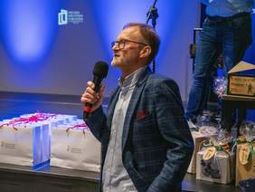 Pokaż zdjęcie: Mężczyzna w okularach, jasnej wzorzystej koszuli i granatowej marynarce w kratę z bordową poszetką przemawia do mikrofonu na scenie. Tło sceny jest intensywnie niebieskie Za mężczyzną stoją rzędy jasnych toreb prezentowych z czerwonymi wstążkami oraz inne zapakowane upominki.