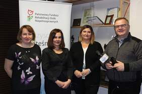 Pokaż zdjęcie: trzy kobiety pracownice Oddziału Lubelskiego PFRON oraz ks. redaktor Wojciech Iwanicki stoją na tle logo PFRON