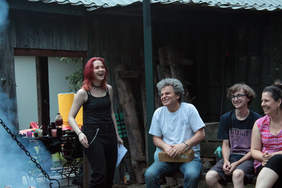 Pokaż zdjęcie: Uśmiechnięta młoda kobieta stoi ubrana w czarną koszulkę. Na ławce obok siedzi uśmiechnięty mężczyzna oraz młoda kobieta i mężczyzna