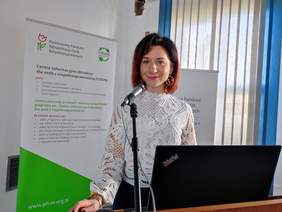 Pokaż zdjęcie: Oparta o mównicę stoi młoda uśmiechnięta kobieta, ubrana w jasną bluzkę.