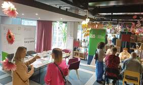 Pokaż zdjęcie: Kobieta stojąc przy stoisku Oddziału Wielkopolskiego PFRON udziela informacji innej kobiecie. Obok znajduje się stolik, a na nim materiały informacyjne. W tle banery PFRON oraz CIDON, a po prawej stronie stoisko Zakładu Ubezpieczeń Społecznych.