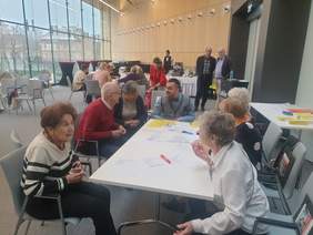 Pokaż zdjęcie: Sala konferencyjna z dużymi oknami. Przy stole siedzi grupa osób pracujących z dokumenta-mi. W tle widoczne są inne osoby i baner „Lublin Gospodarczy”. Zdjęcie przedstawia warszta-ty lub konsultacje.