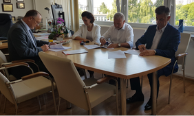 cztery osoby przy stole w biurze, kobieta i trzech mężczyzn, siedzą, pochyleni nad dokumentami, jasny stół, jasne krzesła, z prawej strony okna