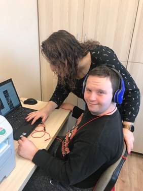 Pokaż zdjęcie: Przy stoliku na który stoi laptop siedzi mężczyzna w niebieskich słuchawkach na uszach nad nim pochylona kobieta.