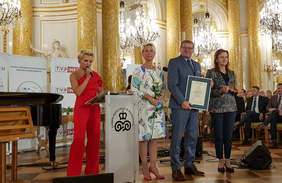 Pokaż zdjęcie: Stojaca przy mównicy kobieta mówi przez mikrofon. Obok mężczyzna trzyma dyplom i kobieta trzymajaca statuetkę.