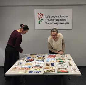 Pokaż zdjęcie: Dwie kobiety pochylone nad stołem z rozłożonymi kartkami świątecznymi wybierające zwycięskie prace.  Na ścianie w środkowej jej części logo Państwowego Funduszu Rehabilitacji Osób Niepełnosprawnych.