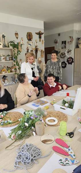 Pokaż zdjęcie: W ośrodku przy stole siedzą trzy osoby z niepełnosprawnościami, wykonują prace ręczne, obok stołu stoją dwie kobiety, które z nimi rozmawiają i przyglądają się ich pracy