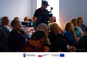 Pokaż zdjęcie: Publiczność siedząca w rzędach na sali podczas wydarzenia słucha wystąpienia. Na środku stoi operator w ciemnym ubraniu i czapce z daszkiem, filmujący zdarzenie kamerą na statywie. Światło wpada z prawej strony, oświetlając profile uczestników. Na dole kadru znajduje się biały pasek z logotypami Funduszy Europejskich, Rzeczypospolitej Polskiej oraz Unii Europejskiej, a w prawym rogu podpis autora: Nico Kaczmarek.