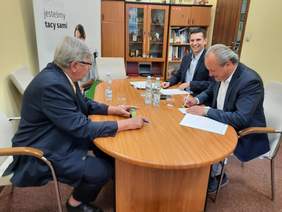 Pokaż zdjęcie: Przy stole siedzi trzech mężczyzn. Uśmiechają się i podpisują umowę. Na stole leżą różne dokumenty, pieczątki, długopisy, woda mineralna. 