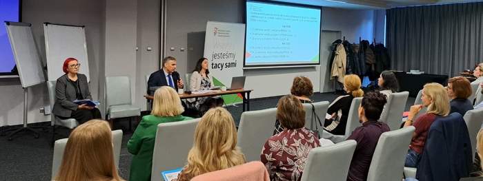 Pokaż zdjęcie: Sala konferencyjna z uczestnikami siedzącymi na krzesłach, przed nimi panel trzech prelegentów przy stole. Ekran z prezentacją, flipcharty, baner z logo PFRO i napisem „Wszyscy jesteśmy tacy sami”, publiczność słuchająca wystąpienia.