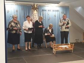 Pokaż zdjęcie: Na scenie stoją cztery starsze osoby z mikrofonami. Jedna z osoba siedzi na krześle. Osoby te trzymają w rękach kartki z tekstem, śpiewają. Wystrój świąteczny, za plecami tych osób na kotarze napis: Bóg się rodzi, naokoło są białe gwiazdki.