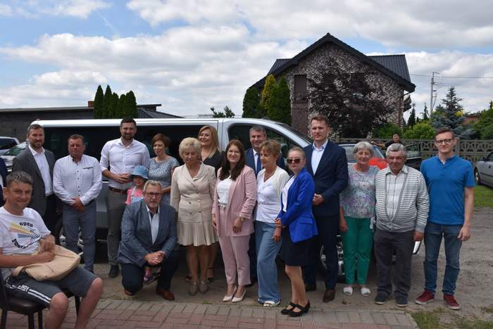 Pokaż zdjęcie: Grupa kilkunastu osób stoi przed srebrnym busem. W tle widoczne są cztery samochody osobowe, dwa budynki mieszkalne oraz drzewa i krzewy. Niebo jest lekko zachmurzone.