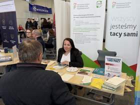Stoisko informacyjne na targach pracy. Kobieta w marynarce rozmawia z mężczyzną siedzącym naprzeciwko przy stole z ulotkami i materiałami. W tle inne stoiska, roll-upy i uczestnicy wydarzenia.