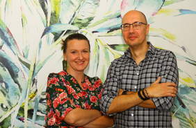 Pokaż zdjęcie: od lewej: uśmiechnięci kobieta i mężczyzna stojący na tle ściany z tapętą w duże liście. Na piersi mają skrzyżowane ręce.