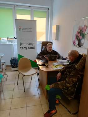 Pokaż zdjęcie: Dwóch mężczyzn siedzi przy biurku, za nimi baner promujący PFRON. Na stole: laptop, ulotki oraz informatory dla osób z niepełnosprawnością.