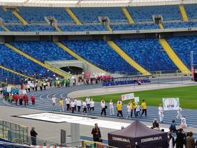 Pokaż zdjęcie: Seniorzy ubrani w sportowe stroje reprezentujący poszczególne UTW maszerują po niebieskiej bieżni stadionu