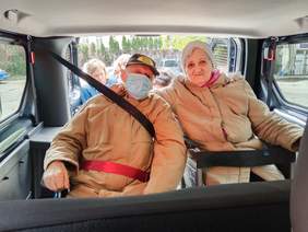 Pokaż zdjęcie: W samochodzie znajduje się starsze małżeństwo. Kobieta obejmuje mężczyznę ramieniem. W tle widać inne osoby. 