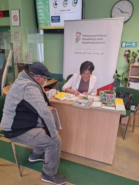 Pokaż zdjęcie: Przy biurku siedzi kobieta oraz mężczyzna, rozmawiają. Na stole leżą ulotki i foldery.