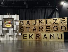 Pokaż zdjęcie: Scenografia teatralna z napisem "BAJKI ZE STAREGO EKRANU" ułożonym z dużych, złotych liter na sześcianach. Po lewej stronie stoi retro telewizor z obrazem testowym. Tło stanowi czarna, zasłonięta kurtyna.