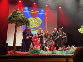 Pokaż zdjęcie: Występujący na scenie aktorzy z niepełnosprawnością, w strojach magów i czarodziejów. W tle tytuł wydarzenia. Po lewej stronie ustawione sztuczne drzewo.