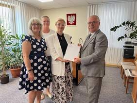 Pokaż zdjęcie: Cztery osoby stoją w biurze podczas oficjalnego podpisania dokumentów. Kobieta w białej marynarce przekazuje teczkę z dokumentem mężczyźnie w szarym garniturze.