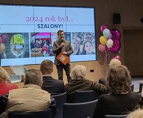 Pokaż zdjęcie: Prelegent prezentuje slajd z napisem „2024 rok był… SZALONY!” oraz zdjęciami z działań Fundacji MOŻESZ w minionym roku. Publiczność uważnie słucha, a po bokach sceny znajdują się balony i dekoracje urodzinowe.