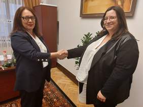 Dwie kobiety o ciemnych włosach, w marynarkach, ściskają sobie dłonie, patrzą w obiektyw aparatu