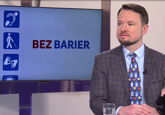 Pokaż zdjęcie: Bliskie ujęcie mężczyzny w średnim wieku z krótkim zarostem, uczestnika programu telewizyjnego. Ma na sobie niebieską koszulę, ciemną marynarkę w kratę oraz niebieski krawat w kolorowe wzory (tulipany). W tle widoczny jest ekran z napisem "BEZ BARIER" oraz piktogramy symbolizujące różne rodzaje niepełnosprawności: ucho (osoby niesłyszące), postać z laską (osoby niewidome) oraz dłonie (język migowy).