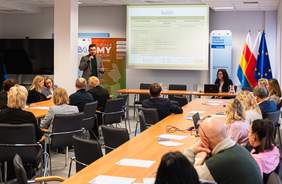 Pokaż zdjęcie: Sala konferencyjna z uczestnikami wysłuchującymi prezentacji o budżecie w ramach projektu „DziałaMY Razem”; prowadzący omawia treści wyświetlane na ekranie.