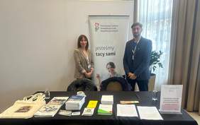 Pokaż zdjęcie: Na zdjęciu kobieta i mężczyzna przedstawiciele PFRON stoją za stołem informacyjnym. Na stole znajdują się ulotki, broszury, torby materiałowe i inne materiały promocyjne oraz tabliczka informująca o Oddziale Podlaskim PFRON. W tle widoczny jest baner PFRON z hasłem „Wszyscy jesteśmy tacy sami”. 