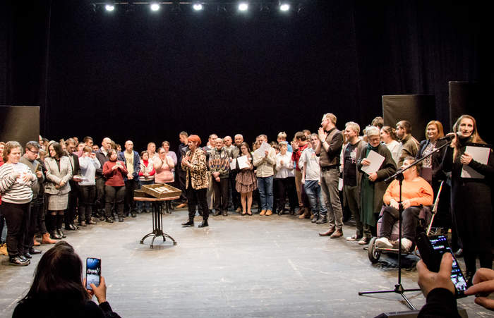 Pokaż zdjęcie: Pamiątkowe zdjęcie wszystkich uczestników festiwalu, którzy stoją na scenie po okręgu. Po prawej stronie kobieta przy mikrofonie wyczytuje laureatów.