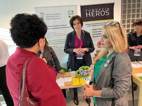 Pokaż zdjęcie: Przed stolikiem dwie kobiety zwrócone do siebie, rozmawiają. Na drugim planie widać stojąca kobietę, z tyłu rollup z logo CIDON.   po