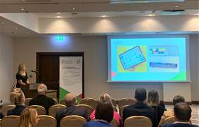 Z lewej strony za mównicą stoi i przemawia kobieta, z prawej wyświetlona na ekranie prezentacja, widać osoby siedzące na krzesłach