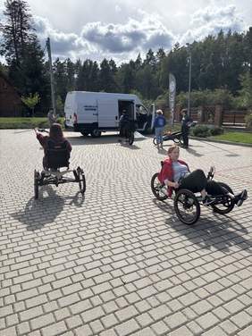 Pokaż zdjęcie: Na placu znajdują się osoby poruszające się na specjalistycznych wózkach oraz rowerach. W tle widoczny jest auto, samochód i drzewa.