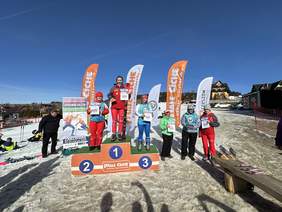 3 osoby, narciarze na podium, z prawej strony poniżej jeszcze trzy, z lewej mężczyzna za nim kilka osób siedzi na śniegu. Na drugim planie banery reklamowe.