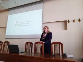 Pokaż zdjęcie: Za stołem konferencyjnym z mikrofonem w ręku stoi kobieta w ciemnym garniturze.