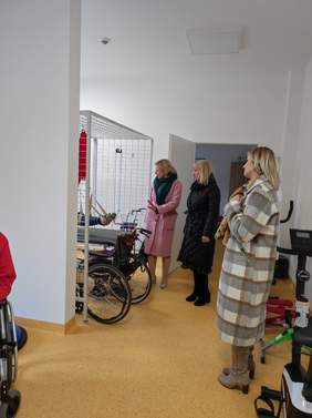 Pokaż zdjęcie: Sala rehabilitacyjna na zdjęciu widać osobę ćwiczącą z podwieszonymi nogami, koło niej stoją trzy kobiety w wierzchnich ubraniach.