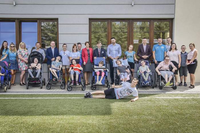 Pokaż zdjęcie: Grupa około trzydziestu osób na tle budynku z dużymi oknami. Wśród tej grupy znajduje się osiem osób z niepełnosprawnością poruszających się na wózku. Z przodu boisko ze sztuczną trawą, na którego skraju leży mężczyzna z kciukiem uniesionym do góry.