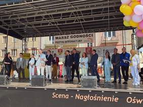Pokaż zdjęcie: Dyrektor Oddziału Podlaskiego oraz przedstawiciele instytucji i organizacji stoją na scenie podczas Festynu Integracyjnego w ramach Marszu Godności Osób z Niepełnosprawnościami w Białymstoku.