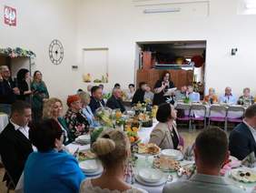 Pokaż zdjęcie: Przy stołach siedzą ludzie. Jedna kobieta stoi i przemawia do mikrofonu. Stoły są zastawione świątecznymi potrawami. 