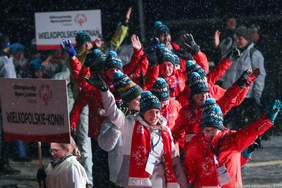 Pokaż zdjęcie: Kilkudziesięcioosobowa grupa osób ubrana w zimowe stroje sportowe macha z radością rękami. Sportowcy trzymają w rękach tabliczki z nazwą miast, które reprezentują i napisem Olimpiady Specjalne